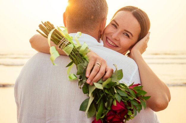 Moglie e marito coppia felice in vacanza al mare. bella donna in abito rosso con un anello al dito e uomo amorevole in una camicia bianca con un mazzo di rose romantico tramonto. Data di San Valentino