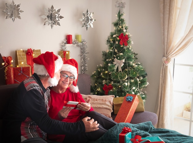 Moglie e marito over 65 seduti sul divano con i cappelli di Babbo Natale che festeggiano il Natale. Un dolce bacio e tanti regali per loro e la famiglia