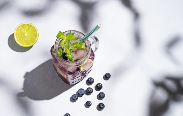 Mojito ai mirtilli o limonata con ghiaccio al lime e menta in un bicchiere su uno sfondo chiaro con frutti di bosco e ombre Il concetto di una bevanda rinfrescante estiva