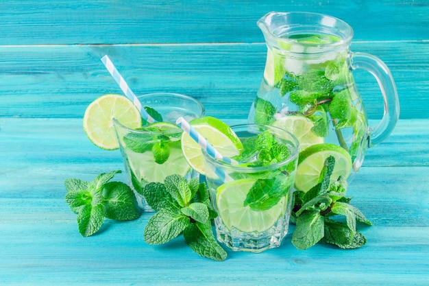 Mojito con menta e lime in un bicchiere e una brocca con tubi. Sfondo di legno blu