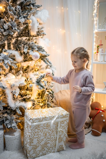 Molto bella e affascinante bambina bionda in pigiama vicino agli alberi di Natale nell'interno luminoso della casa