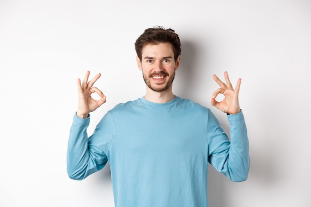 Molto bene. Un bel ragazzo sorridente che mostra segni ok e sembra soddisfatto, in piedi su uno sfondo bianco, approva una scelta eccellente