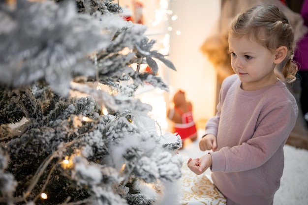 Molto carina e affascinante bambina bionda in pigiama vicino agli alberi di Natale in interni luminosi della casa