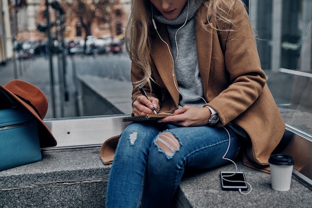 Momenti di ispirazione. Primo piano di una giovane donna che ascolta musica usando il suo smartphone e scrivendo qualcosa mentre trascorre del tempo spensierato in città