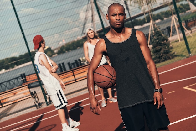 Momento di riposo. Giovane africano in abbigliamento sportivo che guarda l'obbiettivo mentre gioca a basket all'aperto