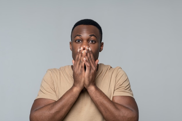 Momento di sorpresa. Sorpreso uomo afroamericano con grandi occhi che coprono la bocca con le mani su sfondo grigio in studio fotografico