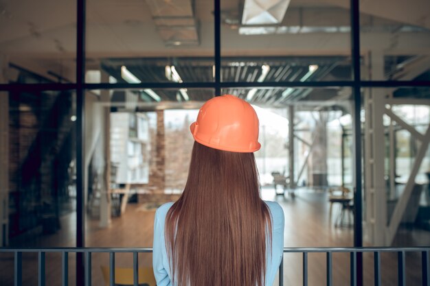 Momento, riflessione. Vista posteriore della donna dai capelli lunghi con casco protettivo e camicetta leggera in piedi con calma nel nuovo edificio pensando