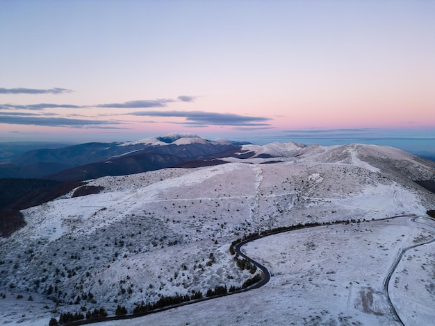 Montagne innevate al tramonto con una strada tortuosa in primo piano vista aerea