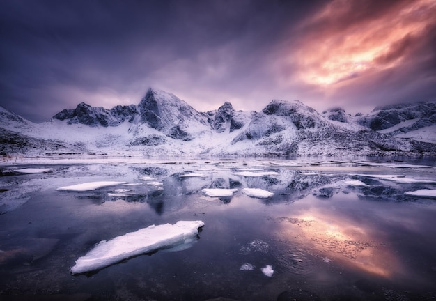 Montagne innevate mare blu con riflesso di ghiaccio costa gelida in acqua e cielo nuvoloso viola al tramonto arancione nelle isole Lofoten Norvegia Paesaggio invernale con fiordo di rocce innevate la sera