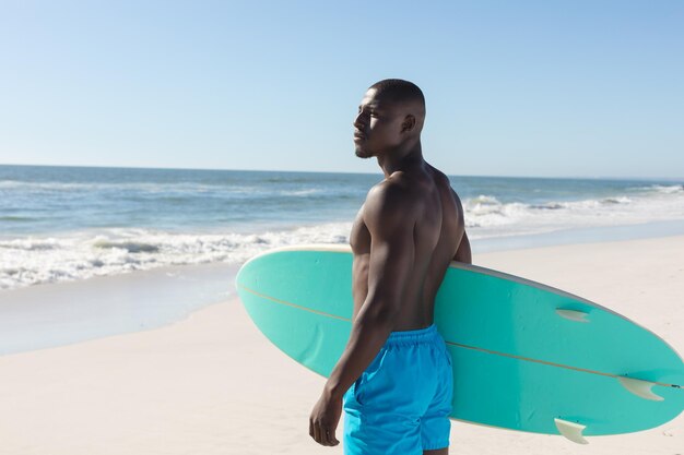 Montare l'uomo afroamericano che tiene la tavola da surf sulla spiaggia soleggiata, distogliendo lo sguardo. Estate, stile di vita sano, sport, hobby, surf e vacanze, inalterati.