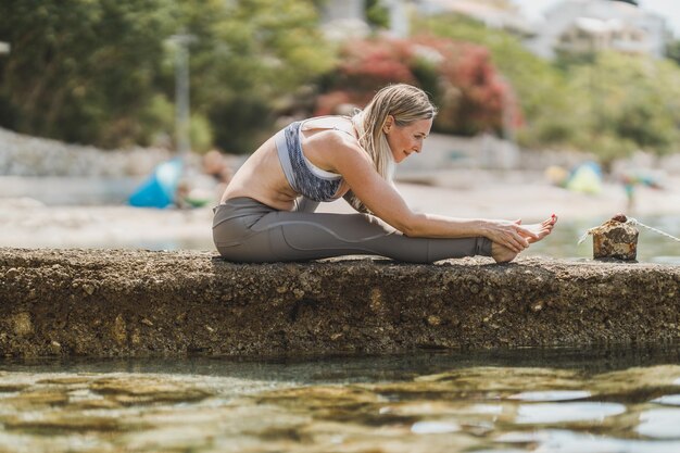 Montare una donna di mezza età che fa esercizi di yoga durante l'allenamento vicino alla spiaggia del mare.