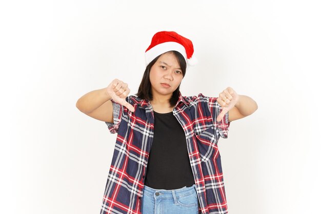 Mostra i pollici in giù della bella donna asiatica che indossa una camicia a quadri rossa e un cappello da Babbo Natale isolato su bianco