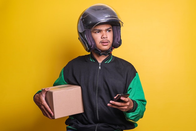 Motocicletta asiatica del tassista online che indossa giacca verde e casco mentre si tiene lo smartphone e il pacco