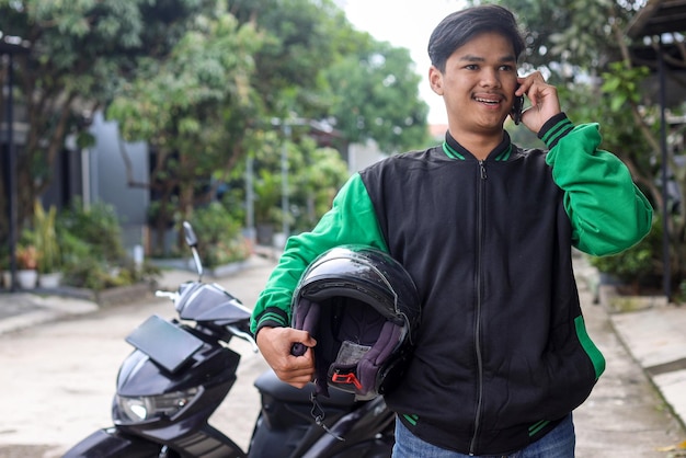 Motocicletta asiatica del tassista online che indossa una giacca verde e tiene il casco con una telefonata