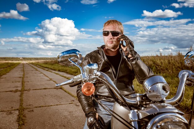 Motociclista che parla su uno smartphone. Uomo del motociclista che indossa una giacca di pelle e occhiali da sole seduto sulla sua moto.