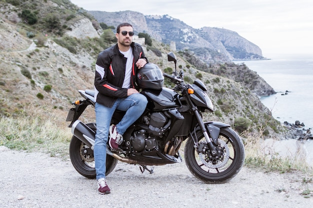 Motociclista del giovane con la sua motocicletta nera pronta a guidare, di fronte al mare