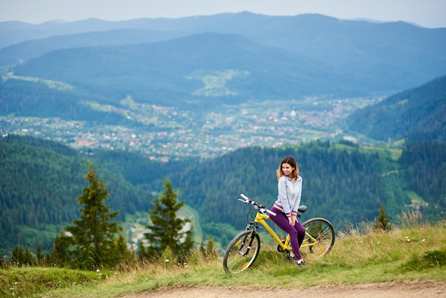 Motociclista della giovane donna che si siede sulla bici gialla vicino alla traccia rurale sulla cima della montagna la sera nuvolosa