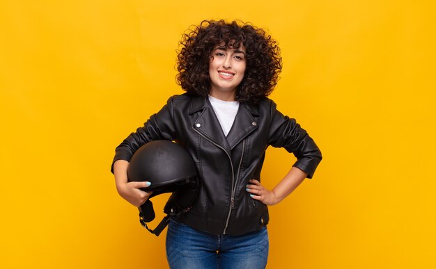 Motociclista donna sorridente felicemente con una mano sul fianco e atteggiamento fiducioso, positivo, orgoglioso e amichevole