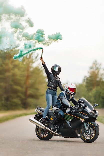 Motociclista in giacca di pelle e casco si siede su una moto sportiva, ragazza in piedi, sfondo sfocato con fumo, copia spazio