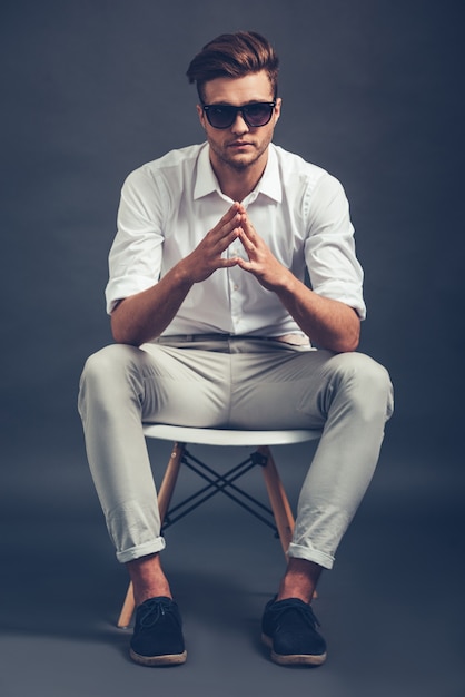 Mr Cool. Tutta la lunghezza di un bel giovane fiducioso in occhiali da sole che tiene le mani giunte e guarda la telecamera mentre è seduto su una sedia su sfondo grigio