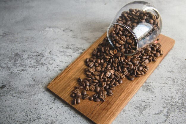 Mucchio di chicchi di caffè in tazza di vetro fuoriuscita su tavola di legno con muro di cemento grigio Freschezza o grande