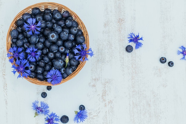 Mucchio di mirtilli freschi e fiori blu in un cesto su fondo di legno bianco