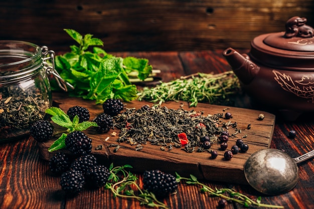 Mucchio di tè verde secco e more fresche sul tagliere di legno. Fasci di foglie di menta e timo. Bollitore di argilla.