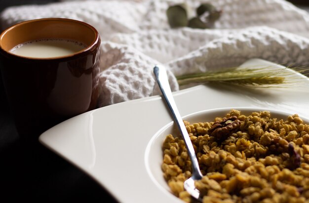 Muesli di cereali in un piatto bianco con un cucchiaio e un bicchiere di latte di argilla su un tavolo in legno nero. messa a fuoco selettiva