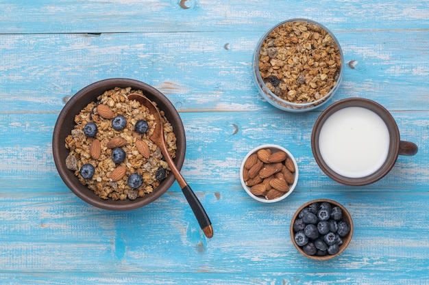 Muesli in una ciotola di argilla, latte, noci e bacche su un tavolo di legno blu. Disposizione piatta.