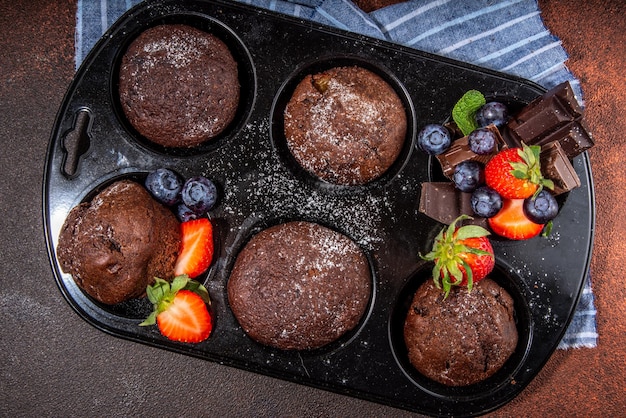 Muffin al cioccolato con frutti di bosco Teglia per muffin con cupcake dolci al cioccolato fondente fatti in casa con salsa di cacao, zucchero in polvere e fragole fresche, mirtilli su sfondo marrone scuro, spazio copia