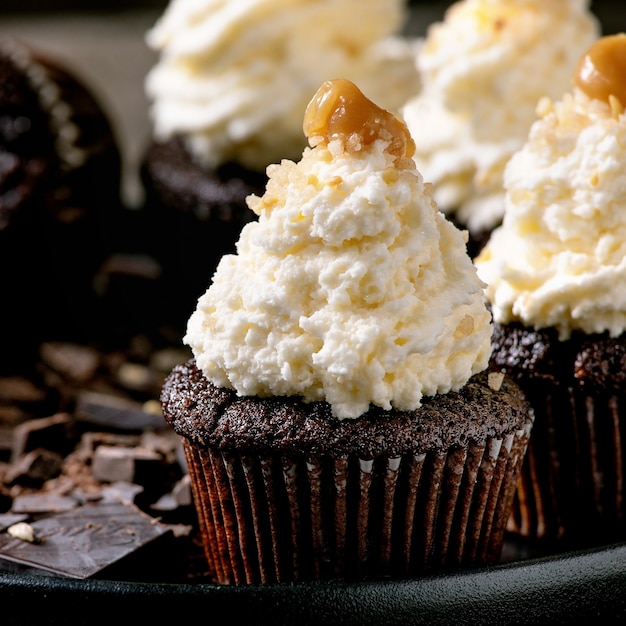 Muffin di cupcakes al cioccolato fatti in casa con crema di burro montata bianca e caramello salato, serviti con cioccolato fondente tritato su piatto di ceramica nera. Avvicinamento. Immagine quadrata