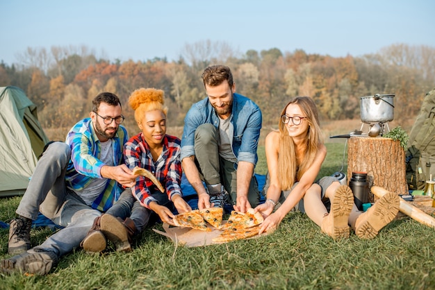 Multi etnico gruppo di amici vestiti casualmente divertendosi a mangiare la pizza durante la ricreazione all'aperto al campeggio