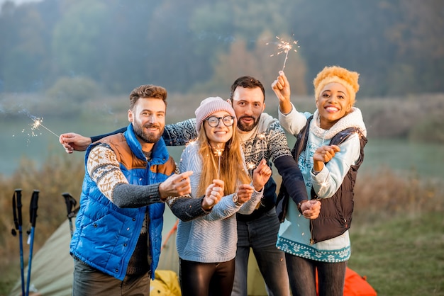 Multi etnico gruppo di amici vestiti con maglioni che celebrano con il fuoco del Bengala in piedi insieme al campeggio durante la luce della sera