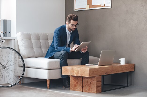 Multitasking. Bel giovane che indossa gli occhiali e lavora con il touchpad mentre è seduto sul divano in ufficio