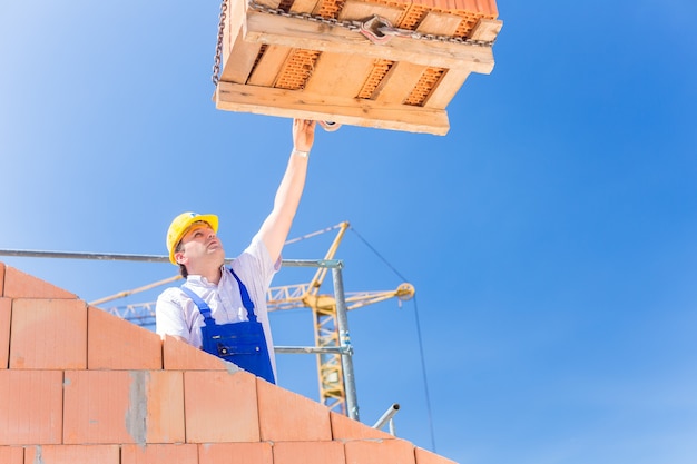 Muratore o operaio edile che riceve una consegna con un pallet di mattoni o pietre dal gruista in costruzione o in cantiere per muri