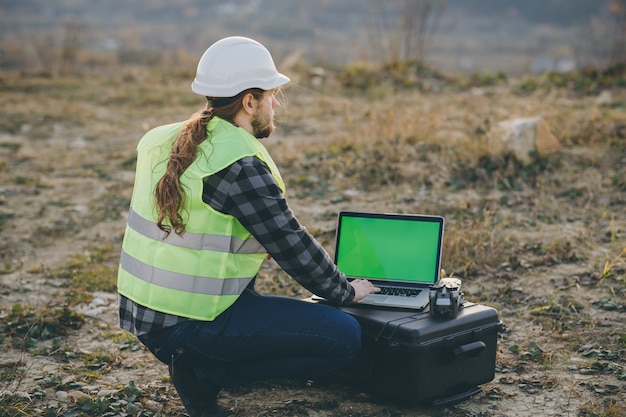 Muratore, uomo caucasico che lavora con il casco di sicurezza e che per mezzo del computer portatile con l'esposizione di chiave dell'intensità