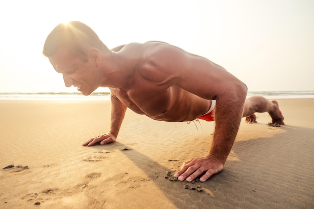 Muscoloso e in forma giovane bodybuilder uomo che fa allenamento warm up stretching su una spiaggia paradisiaca al tramontofitness modello maschile sport sul mare