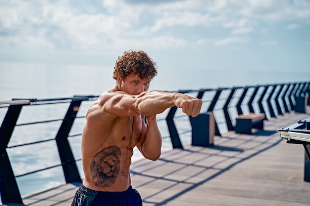 Muscoloso giovane atleta in piedi e praticando shadow boxing all'aperto la mattina presto su pi...