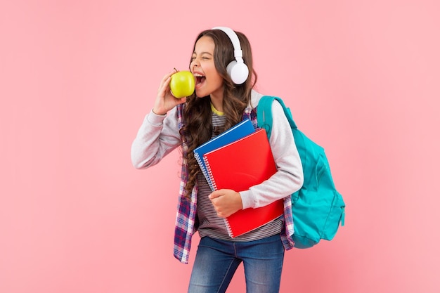 Musica d'ascolto del bambino felice in auricolari con la borsa di scuola e il pranzo della mela del morso della cartella di lavoro