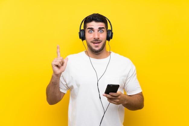 Musica d'ascolto del giovane uomo bello con un cellulare sopra la parete gialla isolata che indica su una grande idea