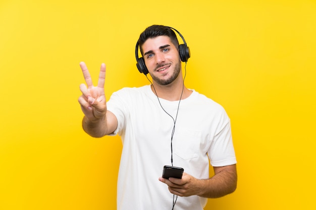 Musica d'ascolto del giovane uomo bello con un cellulare sopra la parete gialla isolata che sorride e che mostra il segno di vittoria