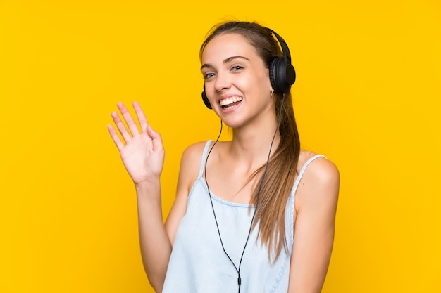 Musica d&#39;ascolto della giovane donna sopra la parete gialla isolata che saluta con la mano con l&#39;espressione felice