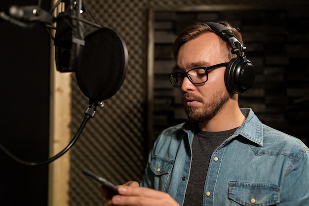 musica, show business, persone e concetto di voce - cantante maschile con cuffie, microfono e smartphone in uno studio di registrazione del suono