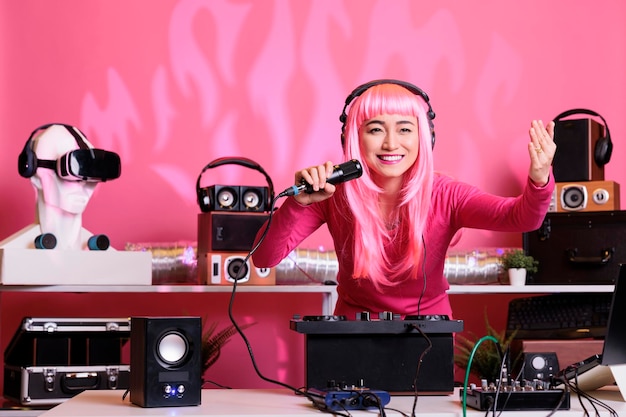 Musicista asiatico seduto al tavolo del dj che esegue musica techno utilizzando giradischi professionali, indossa le cuffie e parla con i fan utilizzando il microfono. Artista con i capelli rosa che suona canzoni elettroniche di notte