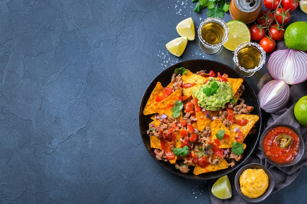 Nachos di tortilla di mais giallo con carne macinata, carne macinata, guacamole, salsa di peperoncino jalapeno rovente e salsa di formaggio con tequila su un tavolo scuro. Vista dall'alto sullo sfondo dello spazio della copia piatta