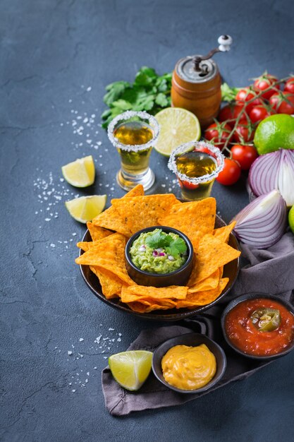 Nachos di tortilla di mais giallo con guacamole, salsa di peperoncino jalapeno rovente e salsa di formaggio con tequila su un tavolo scuro.