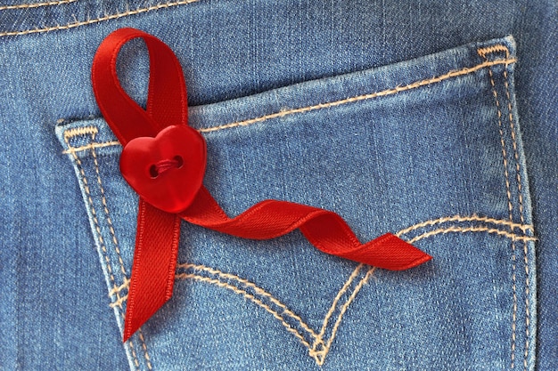 nastro rosso con bottone a forma di cuore sullo sfondo della tasca dei jeans blu - concetto di consapevolezza degli aiuti
