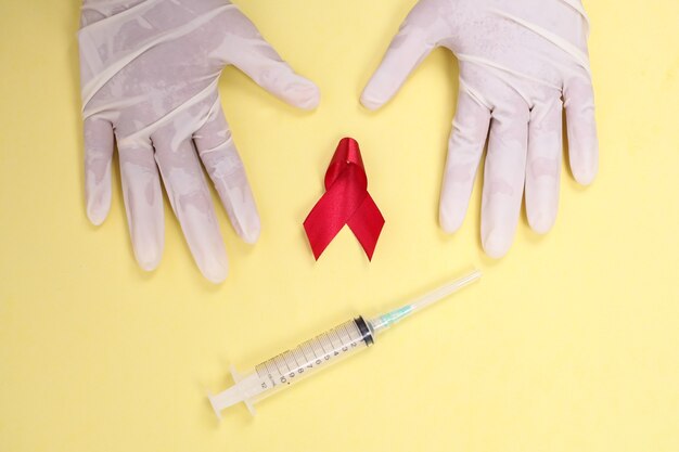 Nastro rosso con guanti medici e siringa contro l'HIV isolato su sfondo giallo