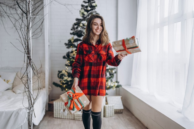 Natale. Bella donna in camicia e calzini che camminano a casa con scatola regalo di Natale, albero di Natale dietro