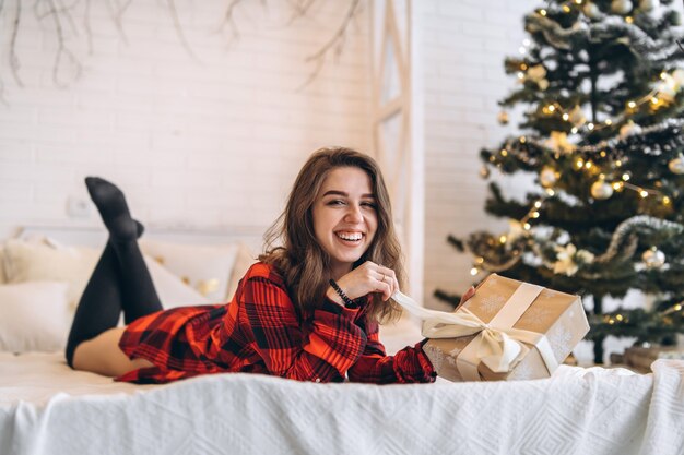 Natale. Bella donna in camicia e calzini sdraiata a letto con scatola regalo di Natale, albero di Natale dietro